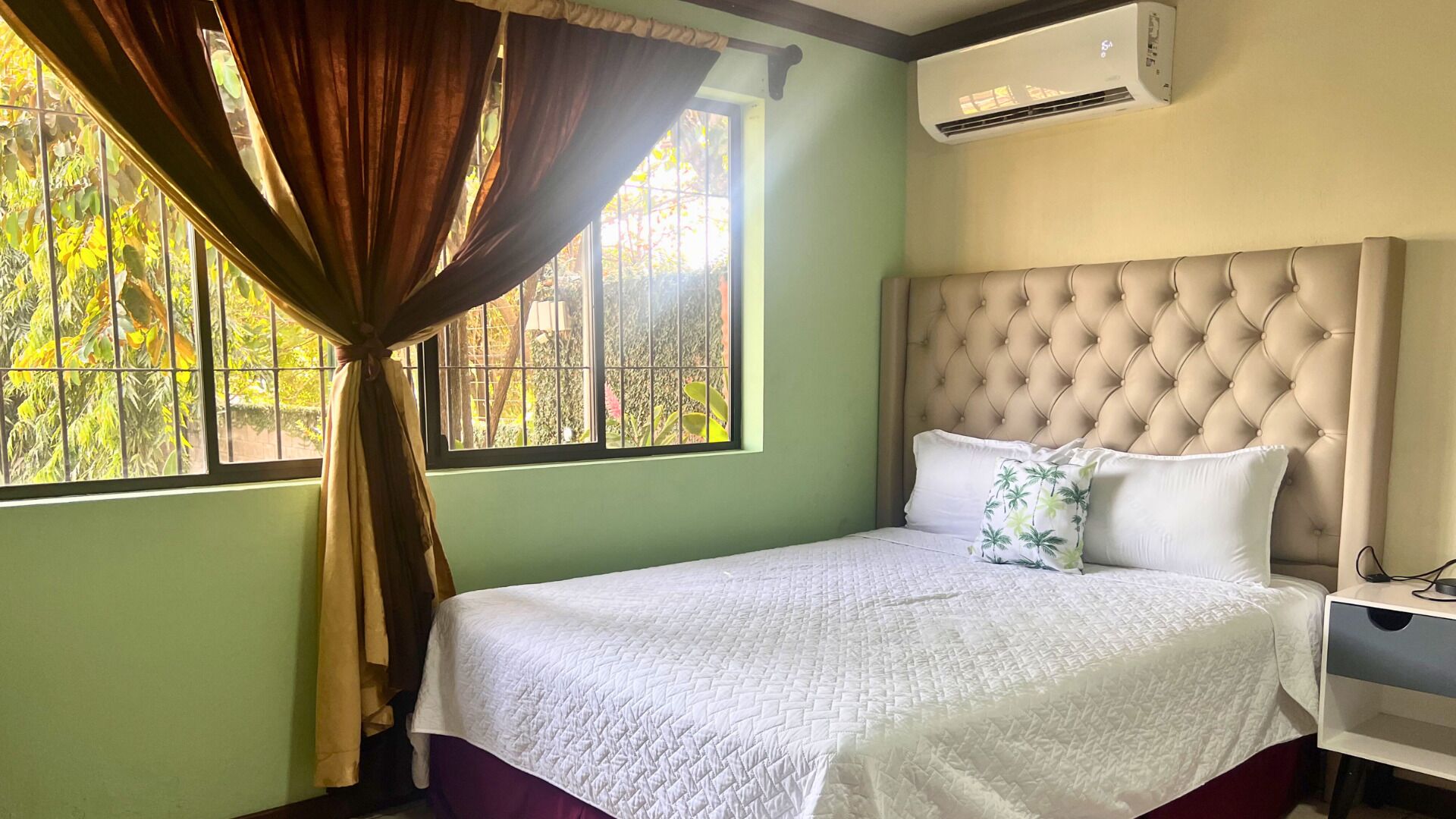 A bedroom with a large window, a tufted headboard, a white bedspread, and a nightstand with a lamp.