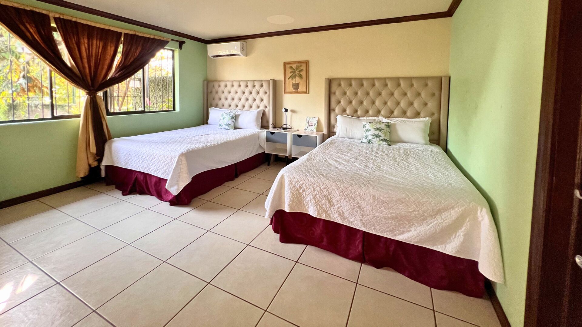 Quarto de hotel com duas camas, uma janela com cortinas, uma mesa de cabeceira e ar condicionado fixado na parede.