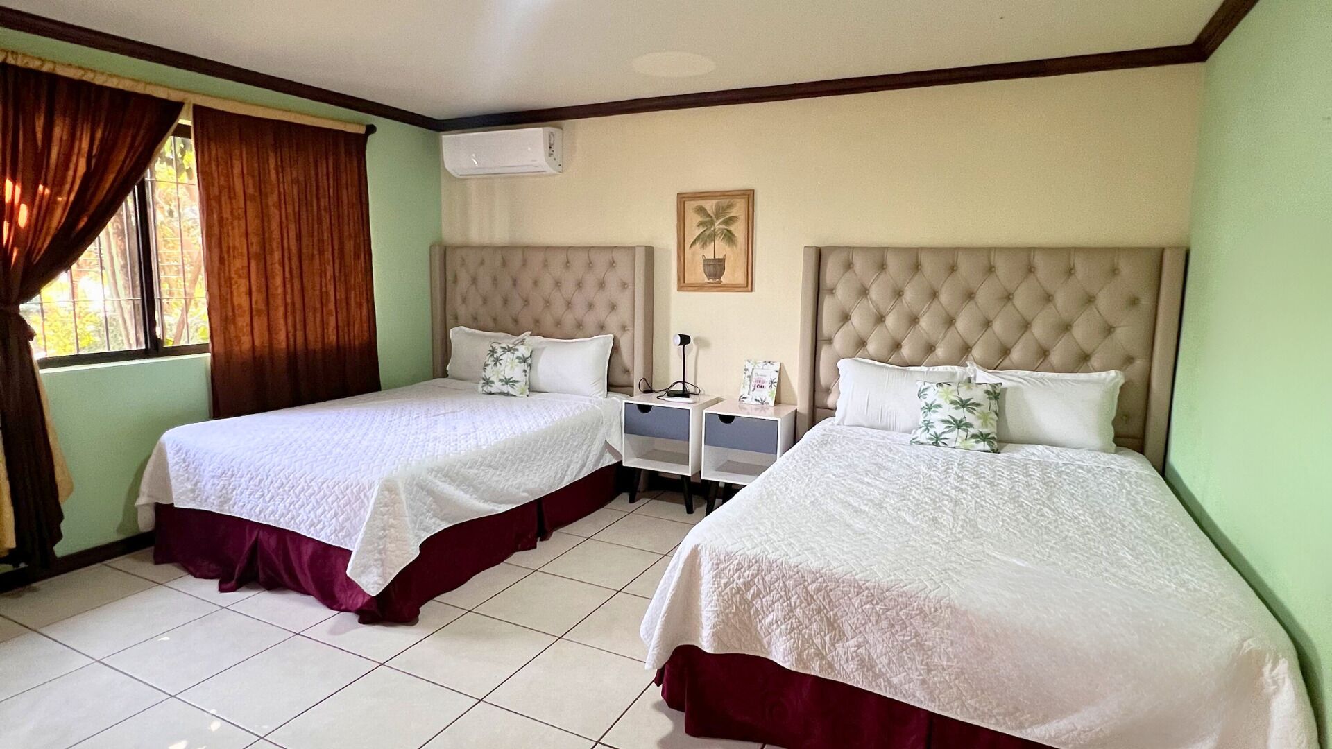 Habitación de hotel con dos camas, cada una con cabecera, una mesita de noche entre ellas y una ventana con cortinas.