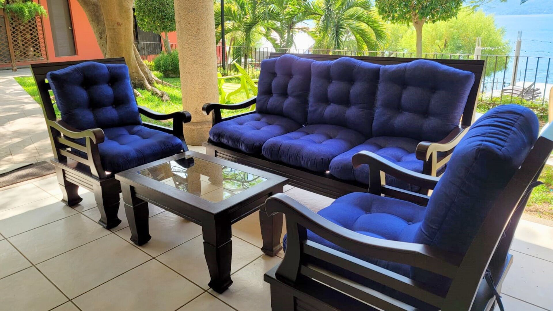 A patio area with a sofa set and chairs, a glass-top coffee table, and a view of a garden and water body.