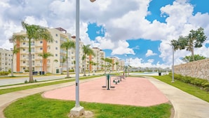 A park with exercise equipment, a paved path, and a building with balconies and palm trees in the background.