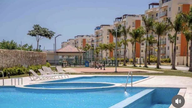 A swimming pool with lounge chairs, surrounded by multi-story buildings with balconies and palm trees.