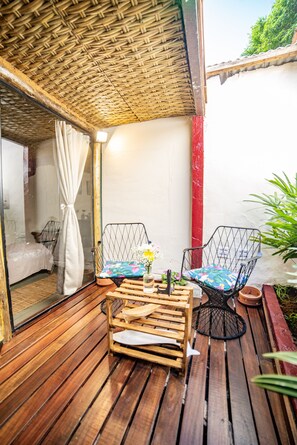A wooden deck with a woven ceiling, a metal chair with a cushion, and a wooden table with a bottle and a flower.