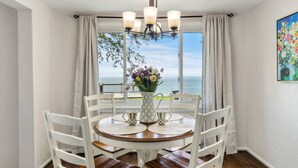 A dining area with a round table, white chairs, a vase with flowers, and a view of the sea through large windows. - Lake Ontario Beauty! Sleeps 4! (Williamson)