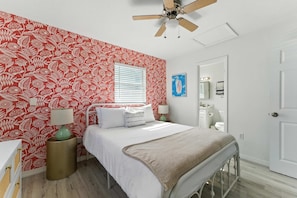 A bedroom with a bed, a bedside table with a lamp, a window with blinds, a wall with a floral pattern, and a ceiling fan.