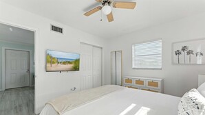 Ein Schlafzimmer mit Bett, Deckenventilator, Fernseher, Fenster mit Jalousien und einer Tür.