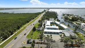 Vue aérienne d’un hôtel situé sur la côte, avec un parking, une route où circulent des véhicules et une marina où sont amarrés des bateaux.