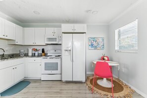 A compact kitchen with white cabinets, a white refrigerator, a microwave, and a stove. There's a dining area with a small table and a red chair.