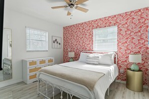 Une chambre à coucher comprenant un lit, des tables de chevet, un ventilateur de plafond et une fenêtre avec des stores.