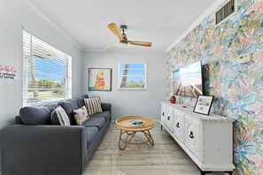 Een woonkamer met een grijze bank, een plafondventilator, een decoratieve wand met tropische behang en een televisie op een wit kastje.