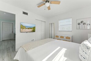 Een slaapkamer met een bed, een plafondventilator, een televisie, een raam met jaloezieën en een deur.