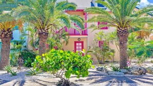 Une propriété située en bord de mer, avec une porte rose, des palmiers et un ciel d’un bleu éclatant.