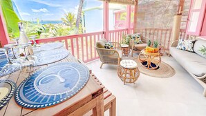 Un espace extérieur abrité, aménagé avec une table en bois, des chaises en osier et une petite table d’appoint en osier sur laquelle sont disposés des fruits.