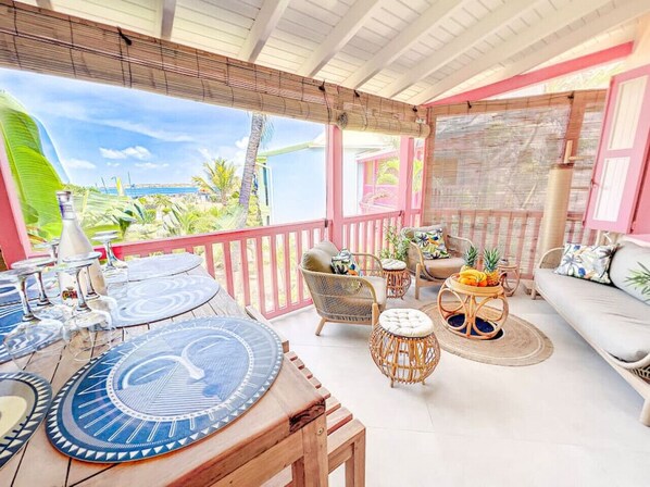 A covered outdoor seating area with a wooden table, wicker chairs, and a wicker side table with fruit. - Costa Rose, spacieux, sur la plage d`Orient Bay (Orient Bay)