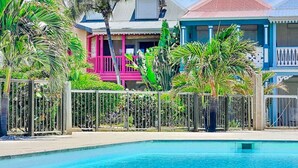 Une piscine entourée d’une clôture, bordée de maisons aux couleurs vives et d’une végétation tropicale.