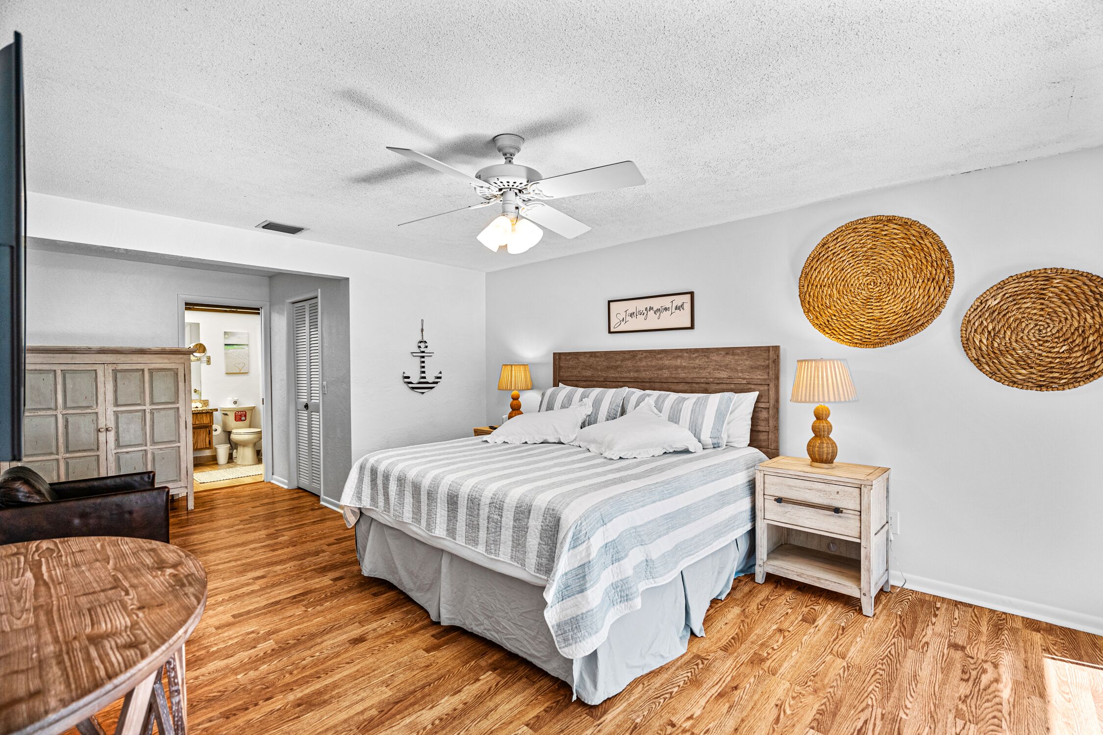 A bedroom with a bed, wooden floor, ceiling fan, bedside lamps, and a wall-mounted decorative item.