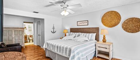 A bedroom with a bed, wooden floor, ceiling fan, bedside lamps, and a wall-mounted decorative item.