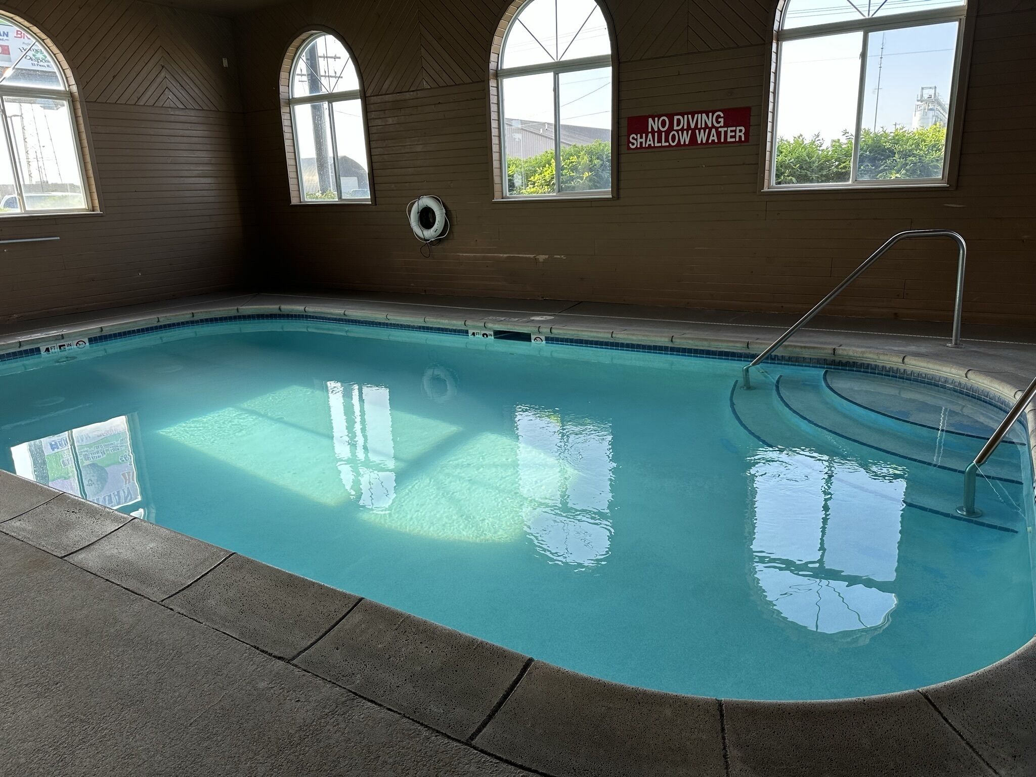 An indoor swimming pool with clear water, a no-diving shallow water sign, and a lifebuoy on the wall.