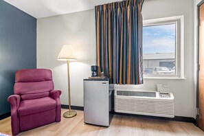 A room with a red chair, a lamp, a small refrigerator, and a window with curtains. - Econo Lodge El Paso, IL (El Paso)