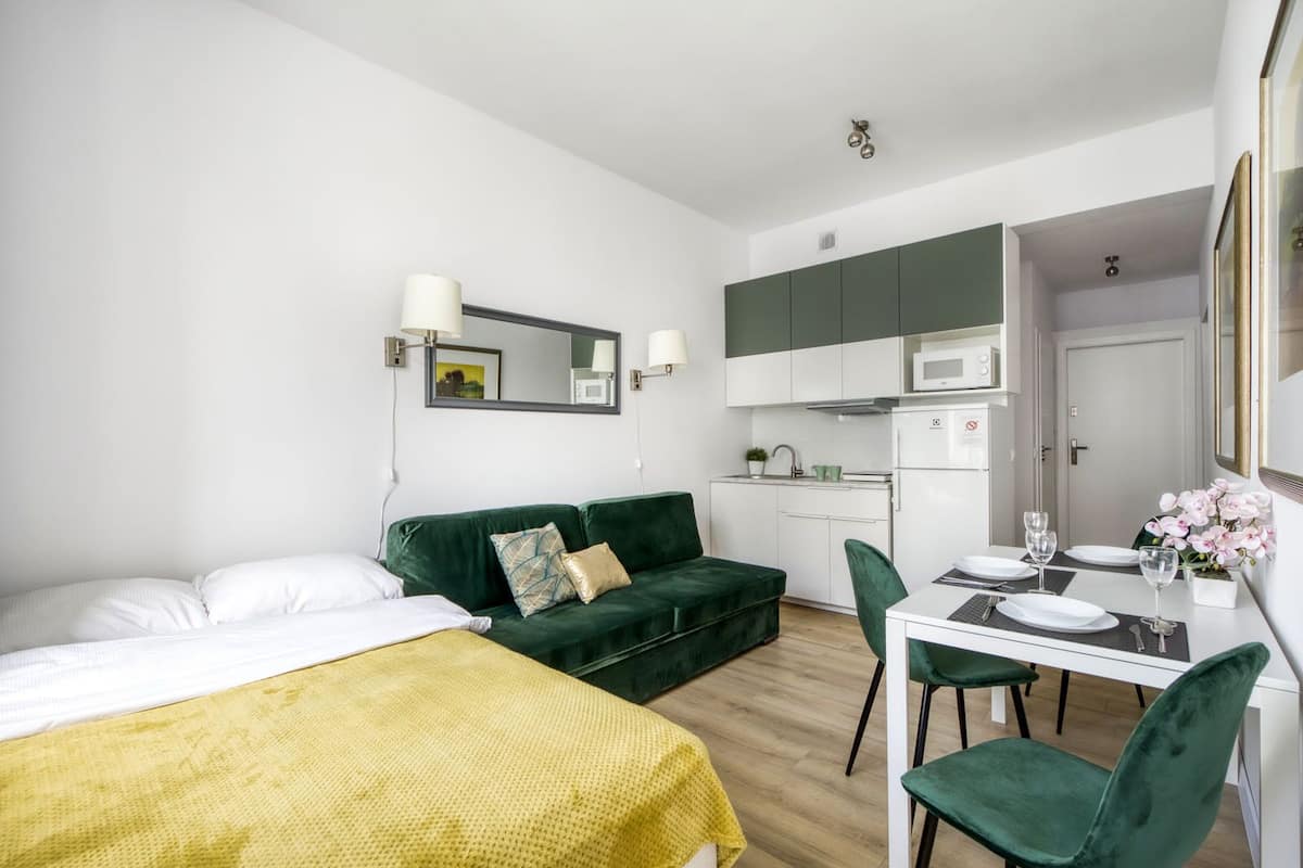 A modern hotel room with a bed, a green sofa, a dining table with chairs, a kitchen area, and a bathroom visible through an open door.