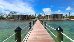 A wooden pier leading to a resort with multiple buildings, surrounded by clear water and palm trees.