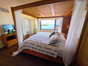 En säng med himmelssäng och utsikt över havet, en tv på ett träställ och en korg av rotting på golvet.