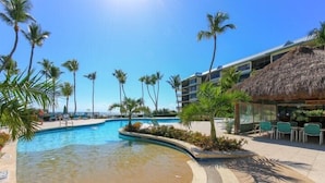 Zona de piscina en un resort con palmeras, una estructura de techo de paja y cielo azul despejado.