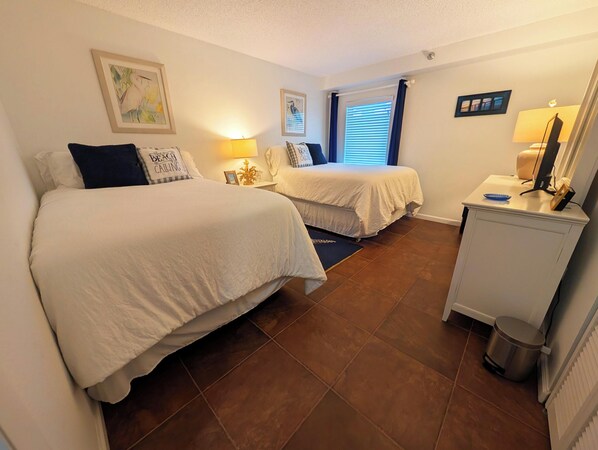 A hotel room with two beds, a desk with a lamp, a trash bin, and framed pictures on the wall.