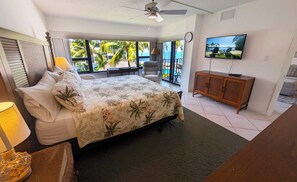 A bedroom with a bed, a television, a wooden cabinet, and a balcony with a view of palm trees and the ocean. - This condo is located in one of Islamorada's most prestigious condo complexes. (Islamorada)