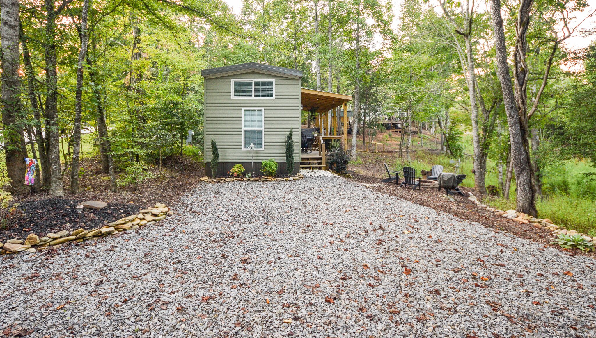 A small cabin with a porch, surrounded by a gravel driveway and wooded area.