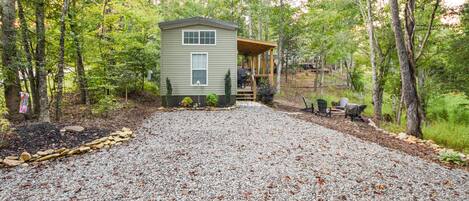 A small cabin with a porch, surrounded by a gravel driveway and wooded area.