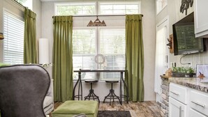 Une pièce chaleureuse avec un fauteuil vert, une petite table avec deux tabourets, une grande fenêtre avec des rideaux et un téléviseur fixé au mur.