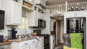Une cuisine moderne avec des armoires blanches, un réfrigérateur noir, un four à micro-ondes et une cuisinière. On y trouve une chaise avec un plaid vert et un coussin.