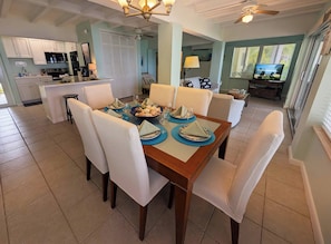 A dining area with a wooden table, white upholstered chairs, and a kitchen in the background.
