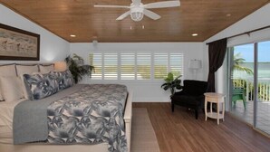 A bedroom with a bed, a ceiling fan, a window with blinds, a chair, a small table, and a view of the outdoors.
