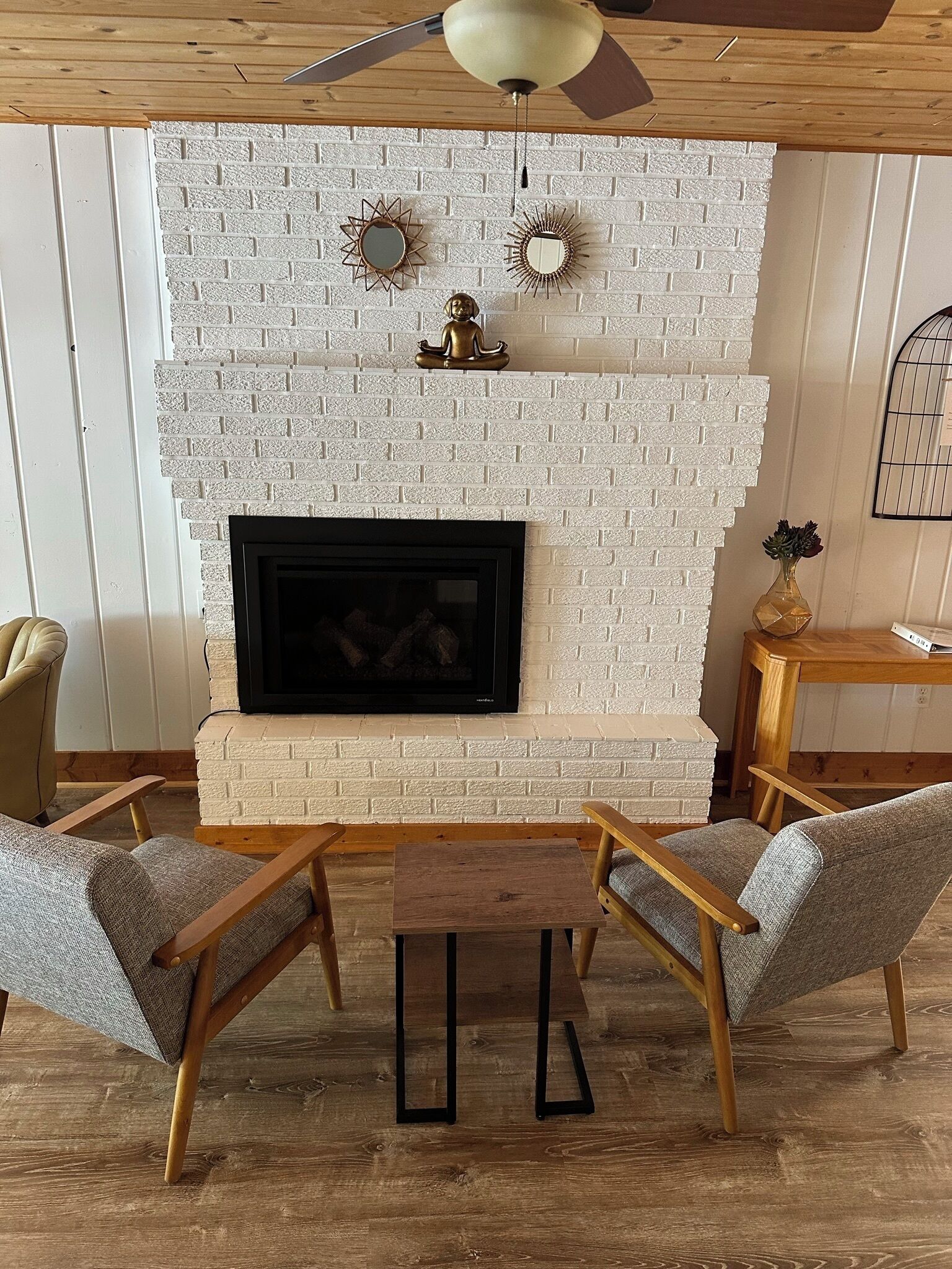 A living room with a white brick fireplace, two armchairs, a small wooden table, and a ceiling fan.