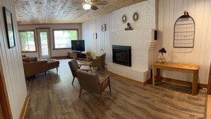 A spacious living room with a fireplace, wooden flooring, and a television. - Loons Landing - Relax on the lake, your perfect Traverse City retreat. (Traverse City)