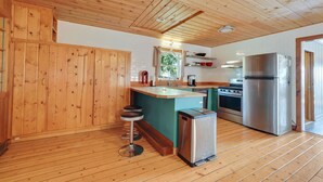 A kitchen with wooden ceiling and walls, a central island with stools, stainless steel appliances, and a white tiled backsplash. - Loons Landing - Relax on the lake, your perfect Traverse City retreat. (Traverse City)