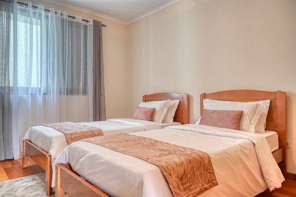 A hotel room with two single beds, a wooden headboard, and a large window with sheer curtains.