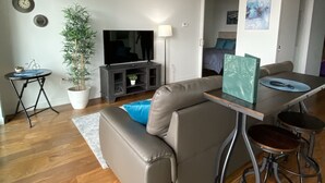 A modern living room with a grey sectional sofa, a small round table, a TV, a potted plant, and a wooden floor. - Downtown luxury 1 bedroom sleeps 3 wifi and laundry  (Winnipeg)