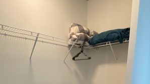Un petit dressing sans porte, avec une étagère en métal, une commode blanche et un couvre-lit bleu.