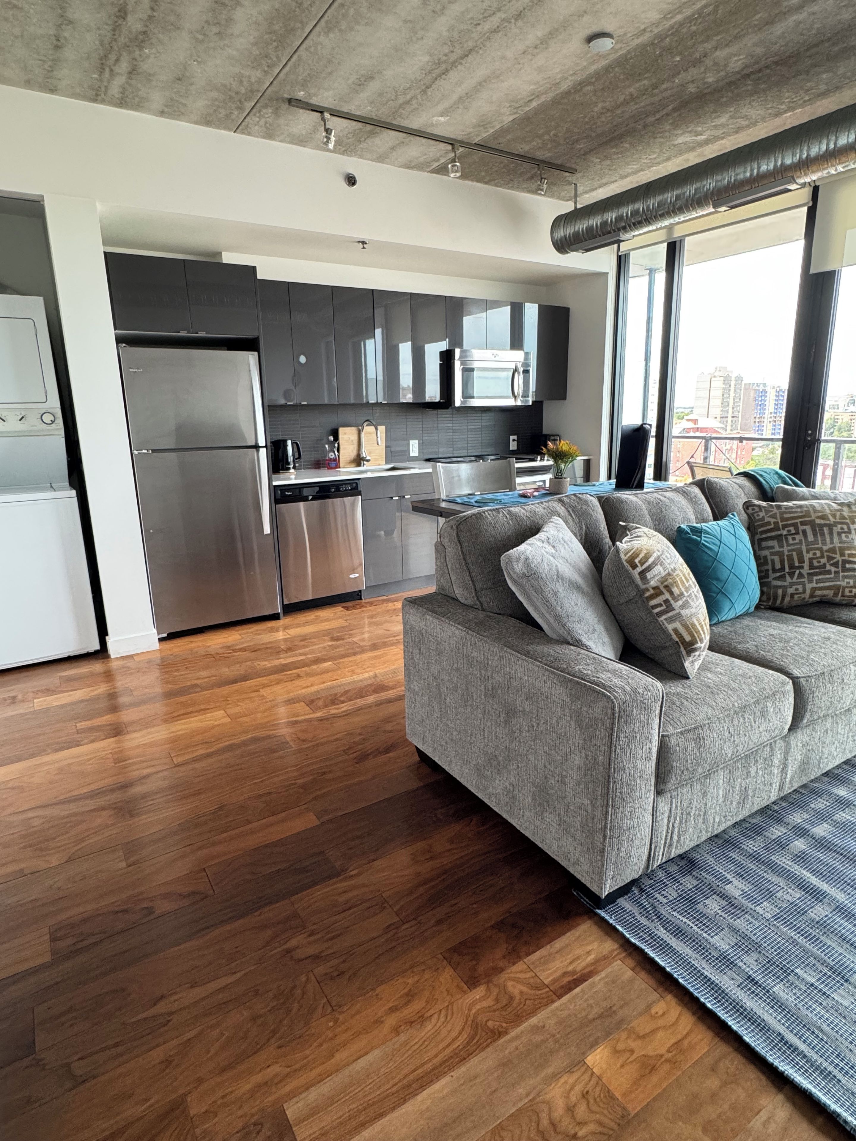 A modern kitchen with stainless steel appliances, a grey sofa with patterned cushions, and a large window offering a view of the city.