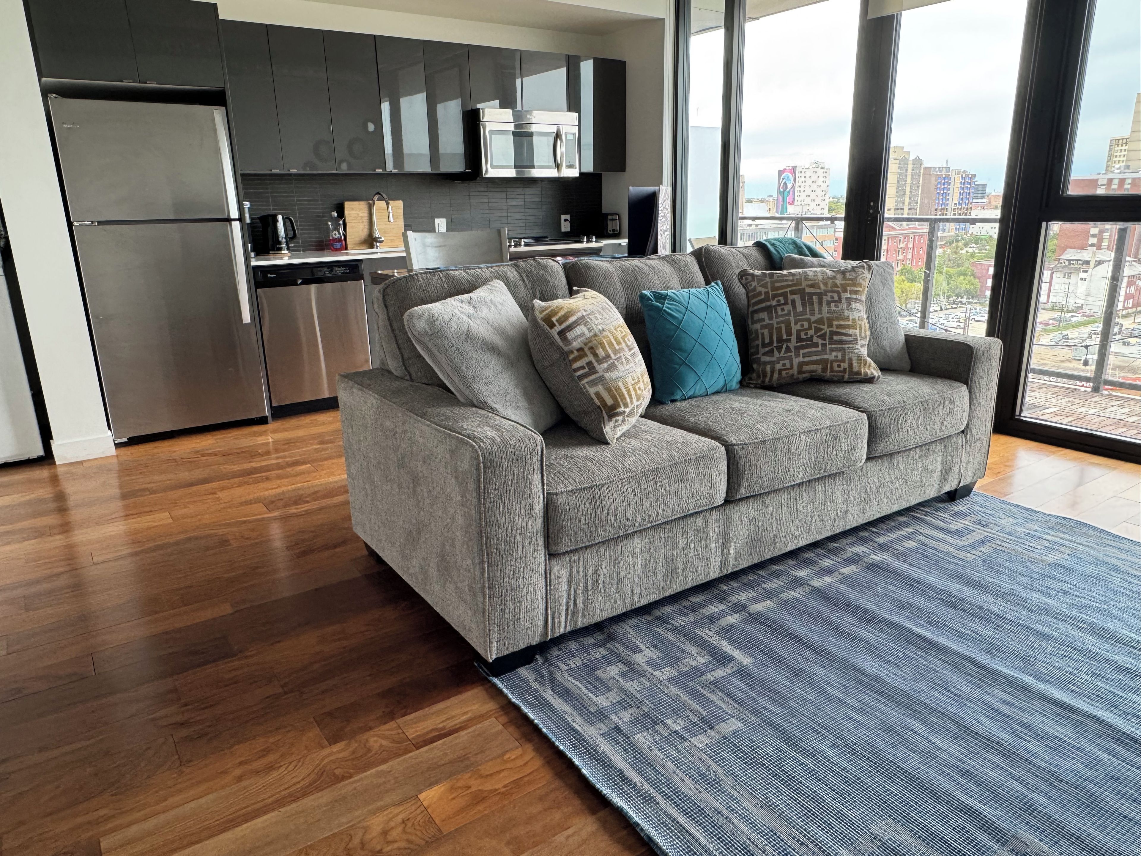 A modern living room with a grey sofa, patterned cushions, and a view of the city through large windows.