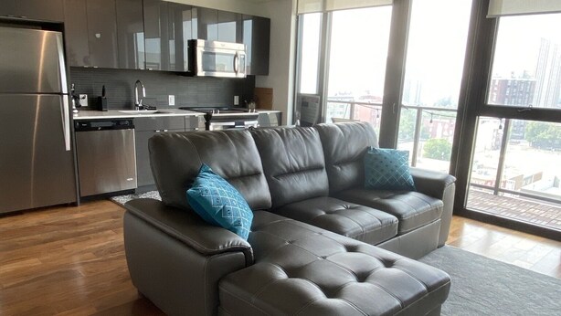 A modern living room with a grey sectional sofa, a kitchen area with stainless steel appliances, and large windows offering a city view. - Downtown luxury 1 bedroom sleeps 3 wifi and laundry  (Winnipeg)
