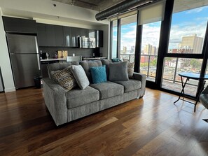 Un espacio habitable moderno con un sofá gris, piso de madera y vistas a la ciudad a través de amplios ventanales.