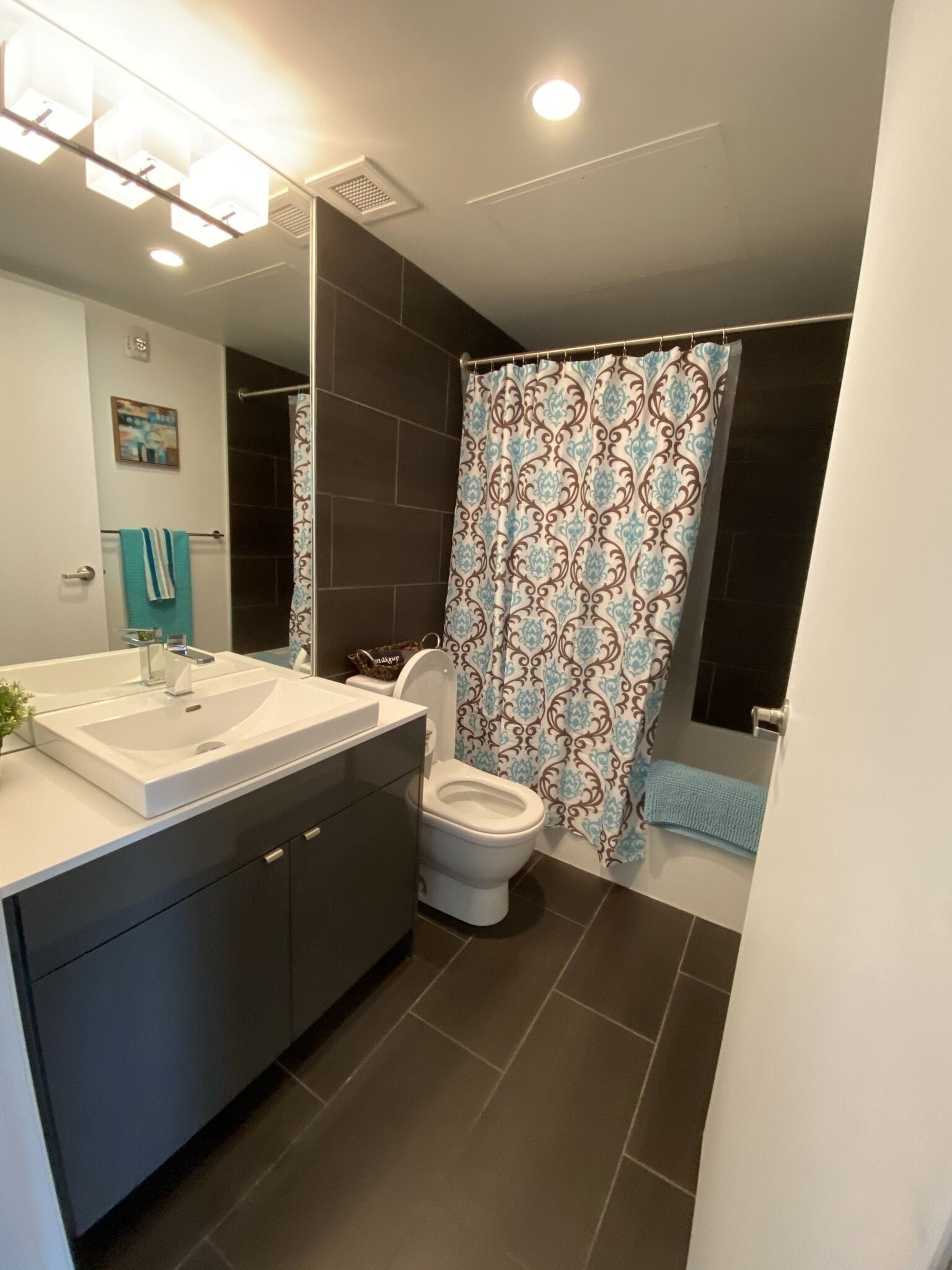 A modern bathroom with a large mirror, white sink, dark cabinets, a toilet, and a shower with patterned curtains.