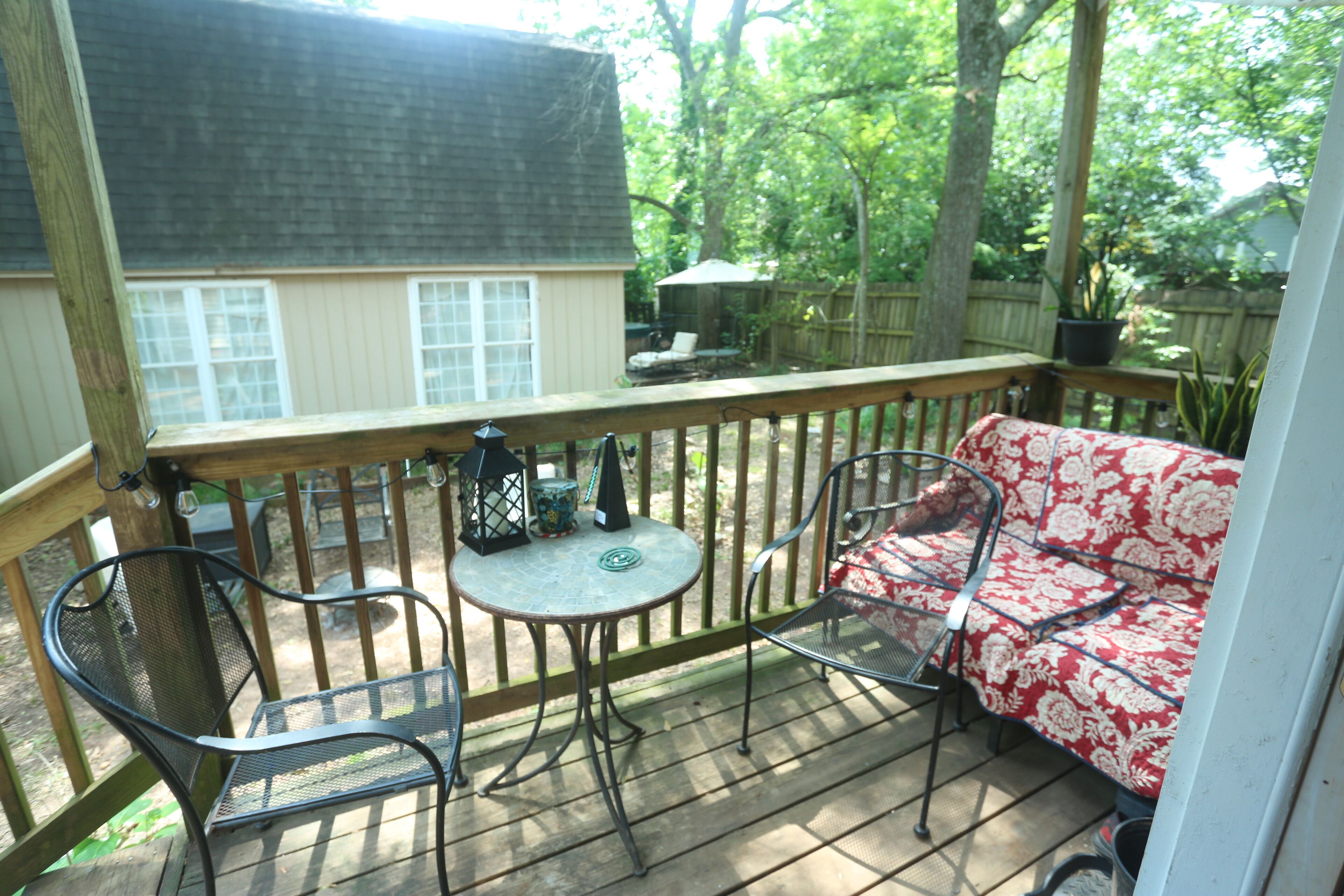 A covered patio with a wooden deck, metal chairs, a small round table, and a red and white patterned sofa.
