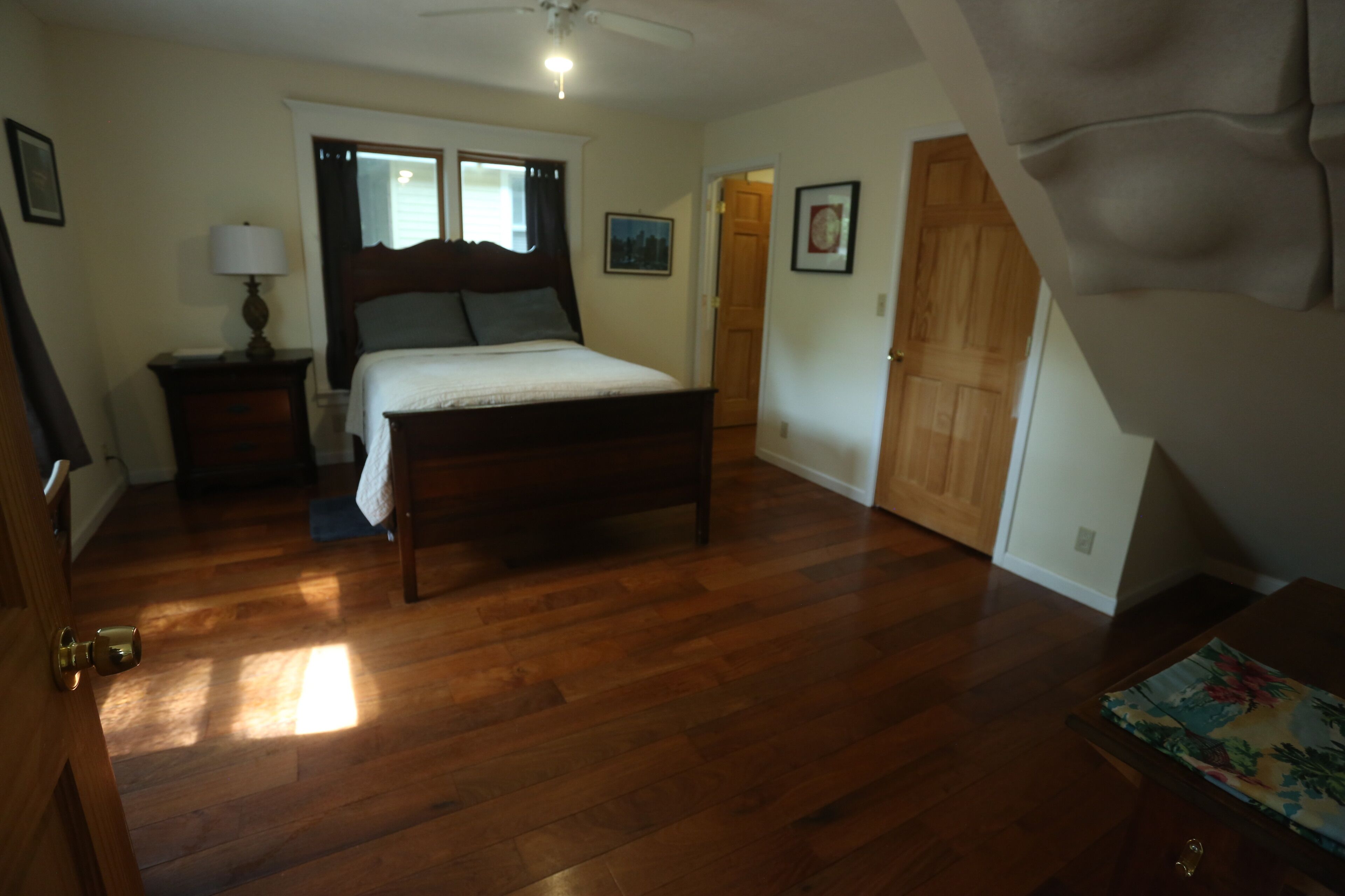 A bedroom with a wooden floor, a bed with a headboard, a nightstand, a lamp, and a door leading to another room.