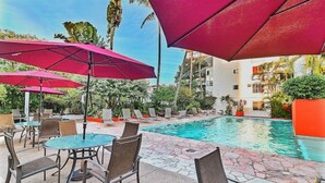 A poolside area with lounge chairs and red umbrellas, surrounded by greenery and a building. - Mexican flair One Bedroom (Puerto Vallarta)