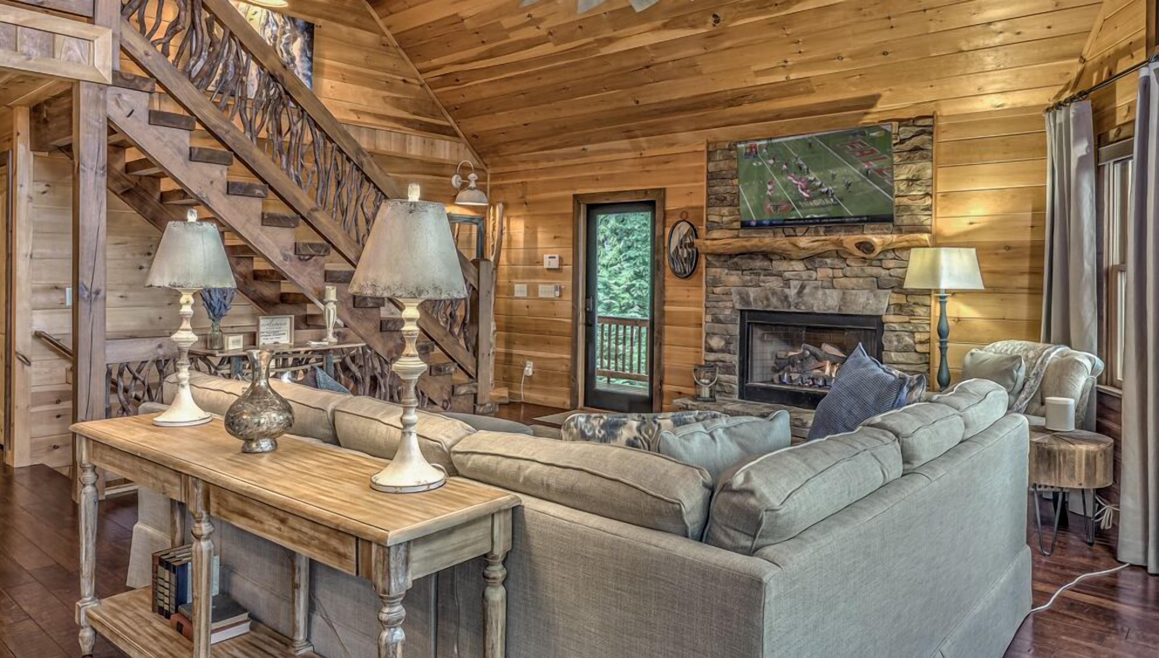 A cozy log cabin interior with a fireplace, a stone wall, a wooden staircase, and a comfortable seating area.